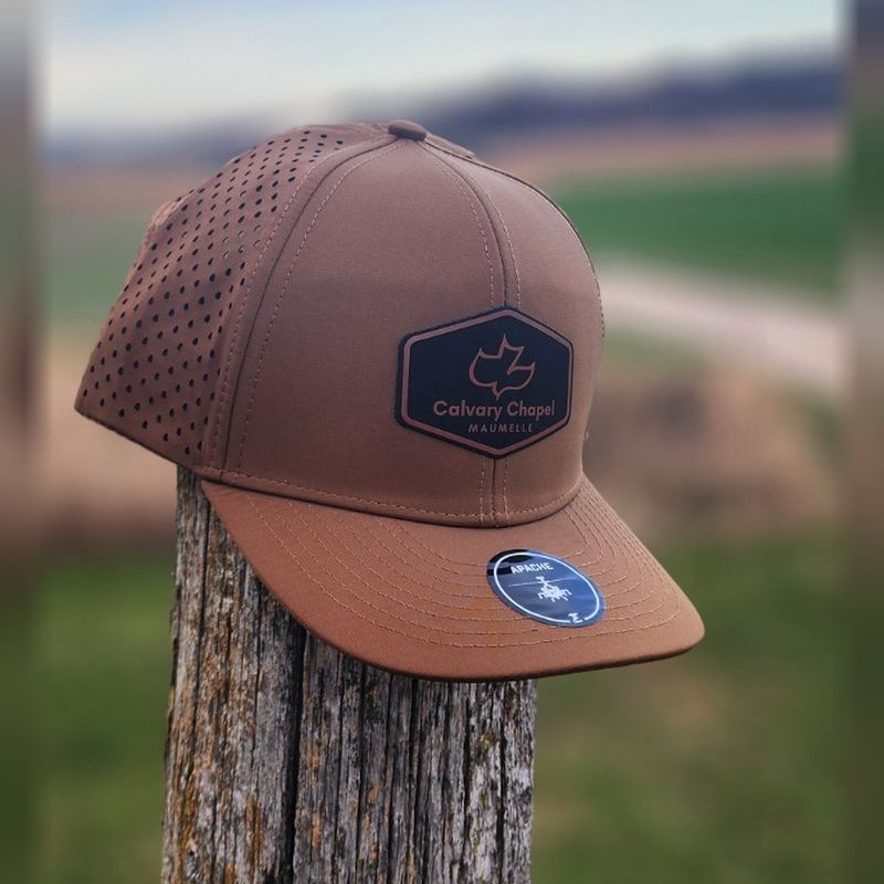 Brown perforated performance hat with a black hexagon Calvary Chapel Maumelle patch, on a fence post