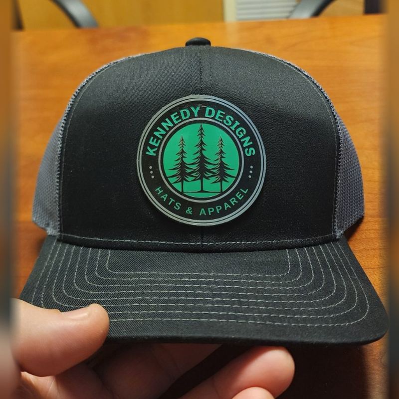 Black trucker hat with a green and black Kennedy Designs pine-tree patch, held in hand