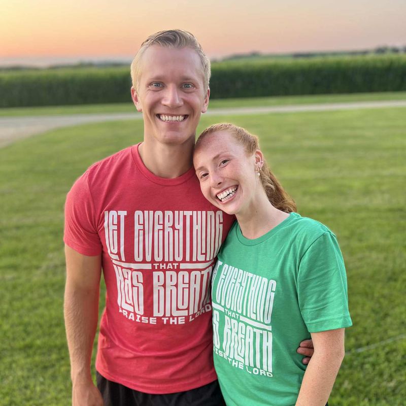 Couple at sunset beside a cornfield wearing red and green heather tees that read 'Let everything that has breath praise the Lord'