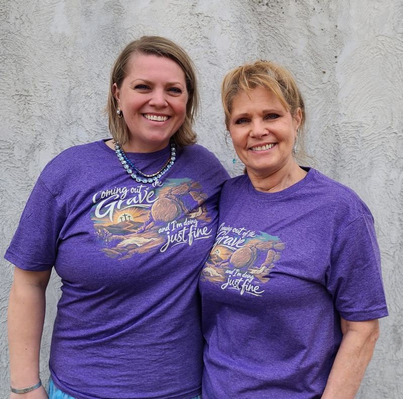 Two women wearing purple heather tees with a full-color 'Coming out of the grave and I'm doing just fine' design
